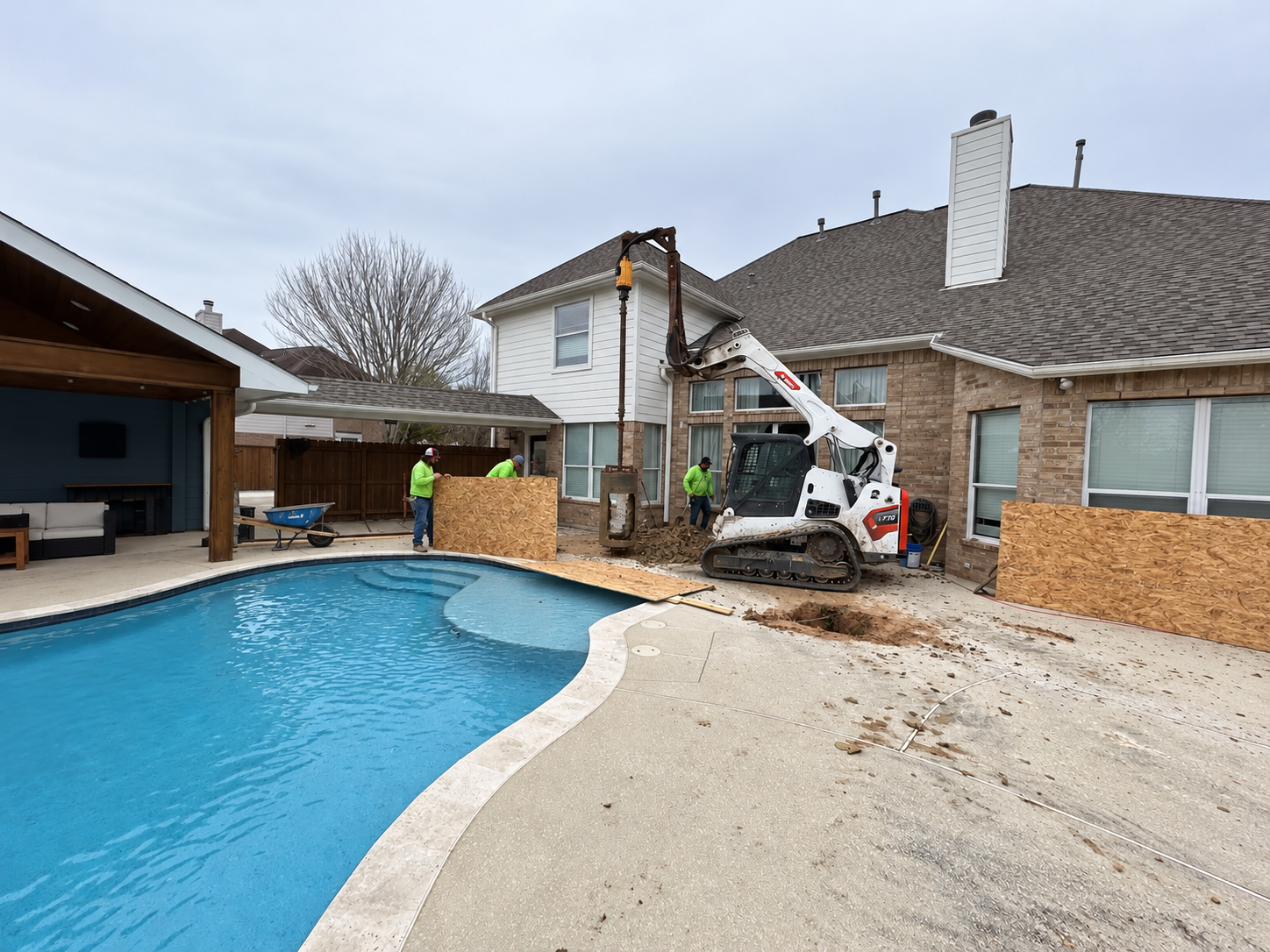 Compact skid steer with a belling attachment in a narrow area beside a pool.