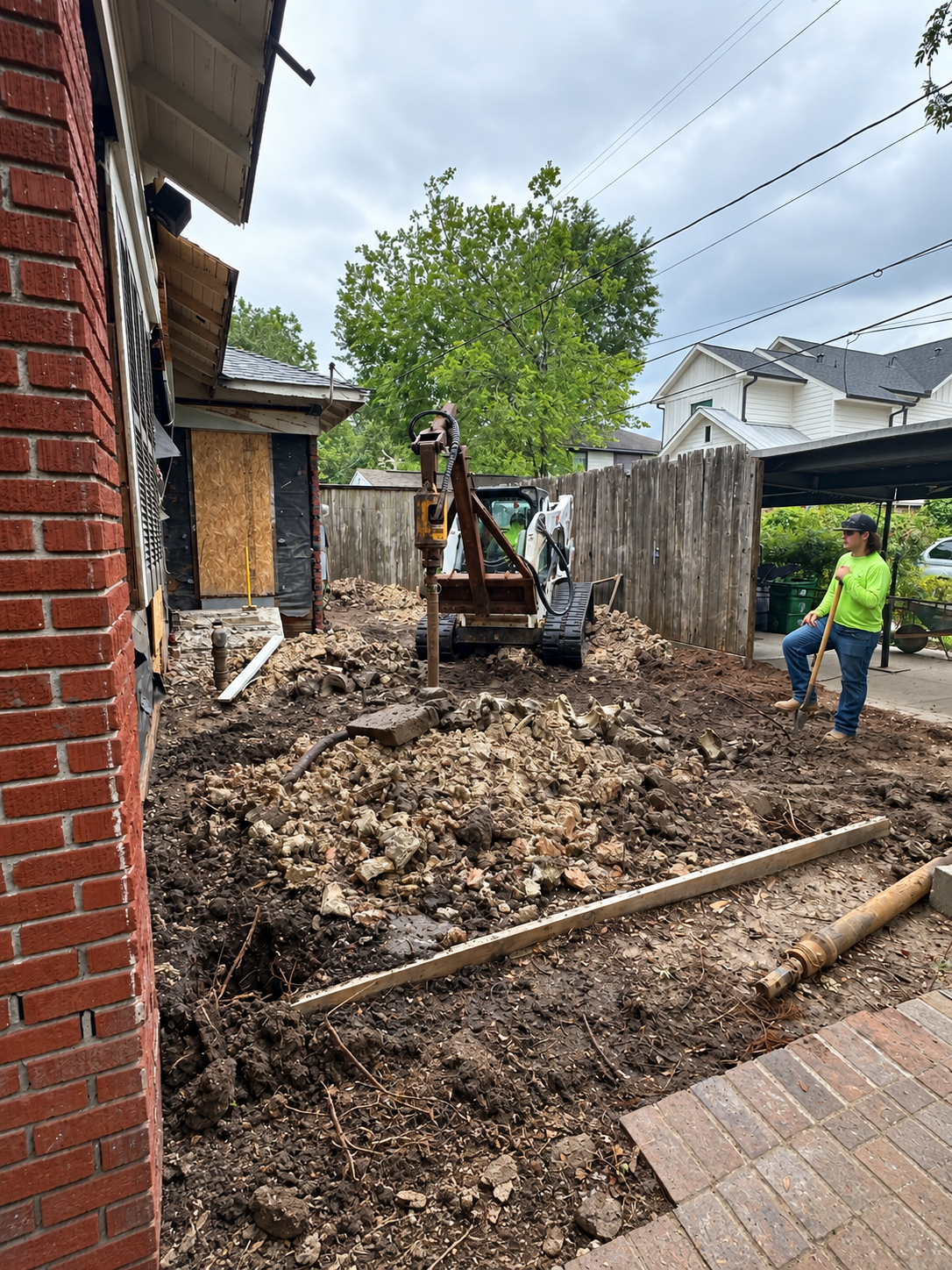 Compact skid steer with a drilling attachment in a narrow residential side yard.