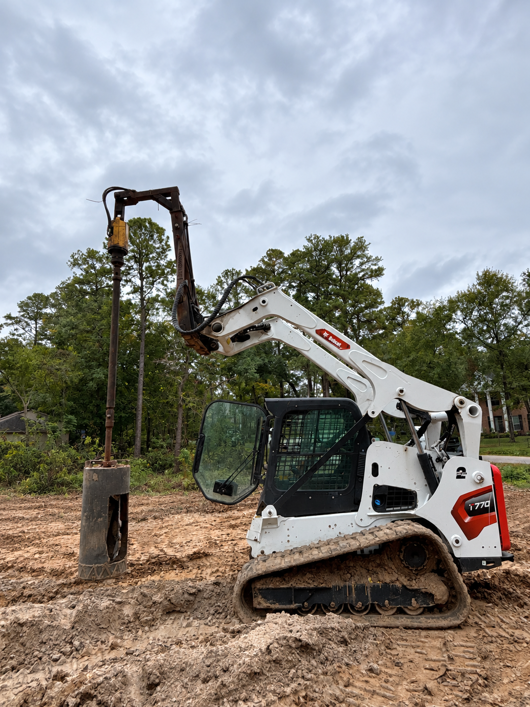 White compact equipment with a vertical auger drilling a pier, with trees behind the machine